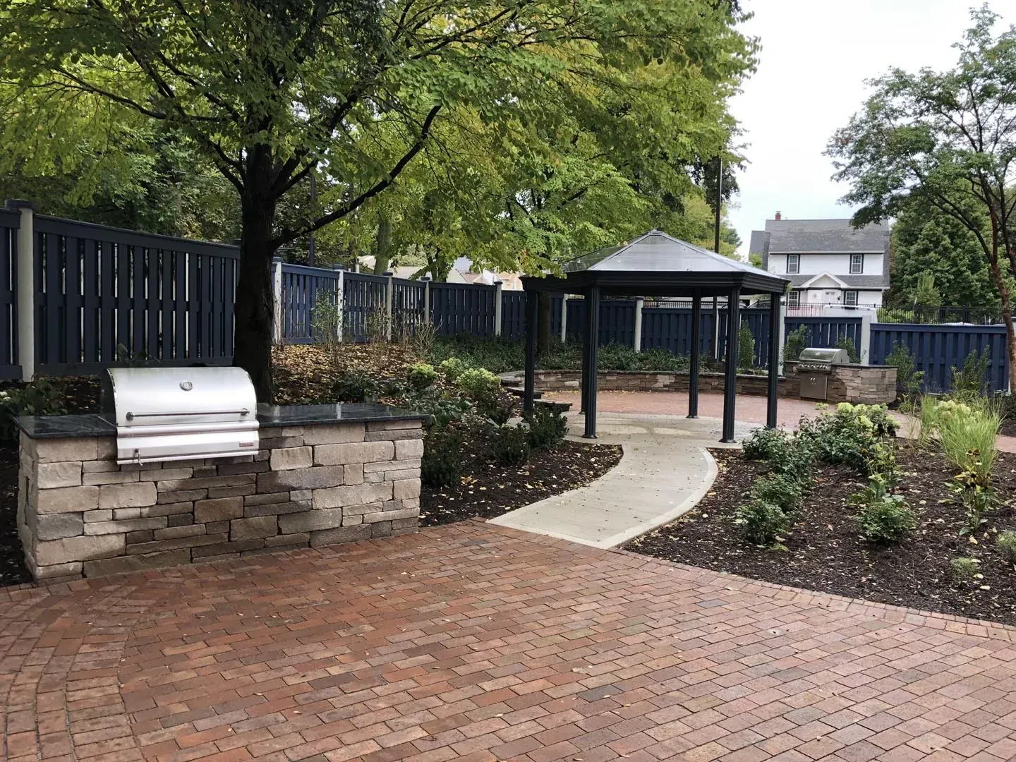 Brick paver patio with an outdoor grill station and a concrete walkway leading to a shaded pavilion