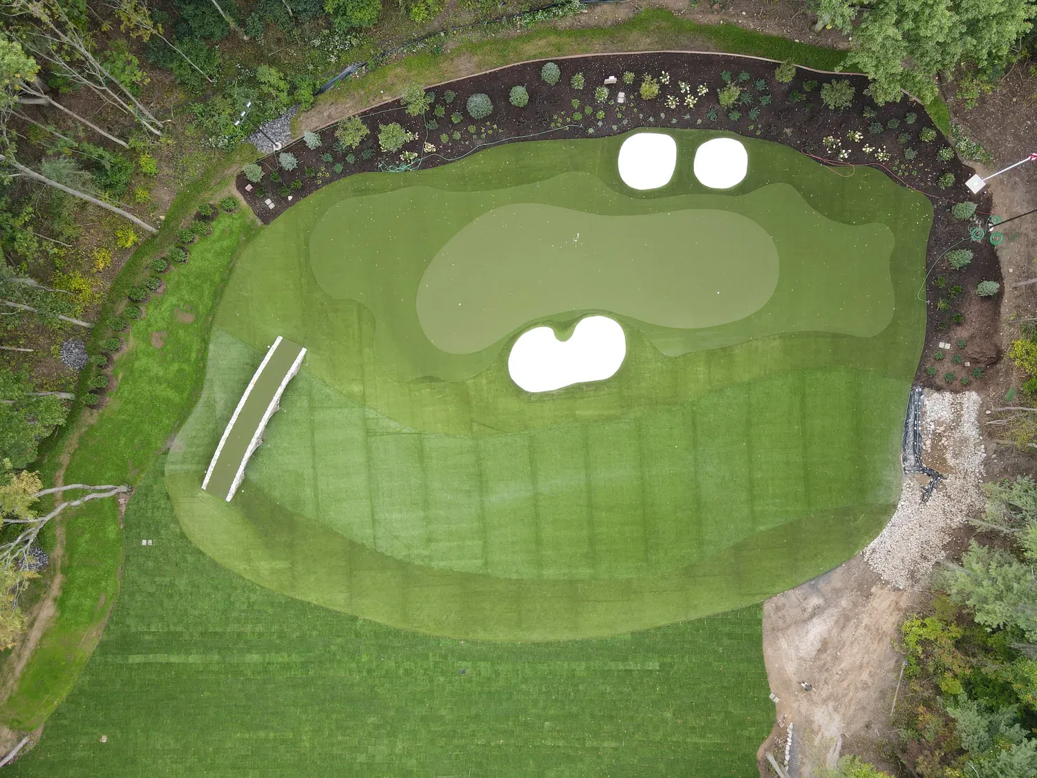 Aerial view of a private artificial-turf putting green with three bunkers, a flag, curved fringe, and a stone bridge