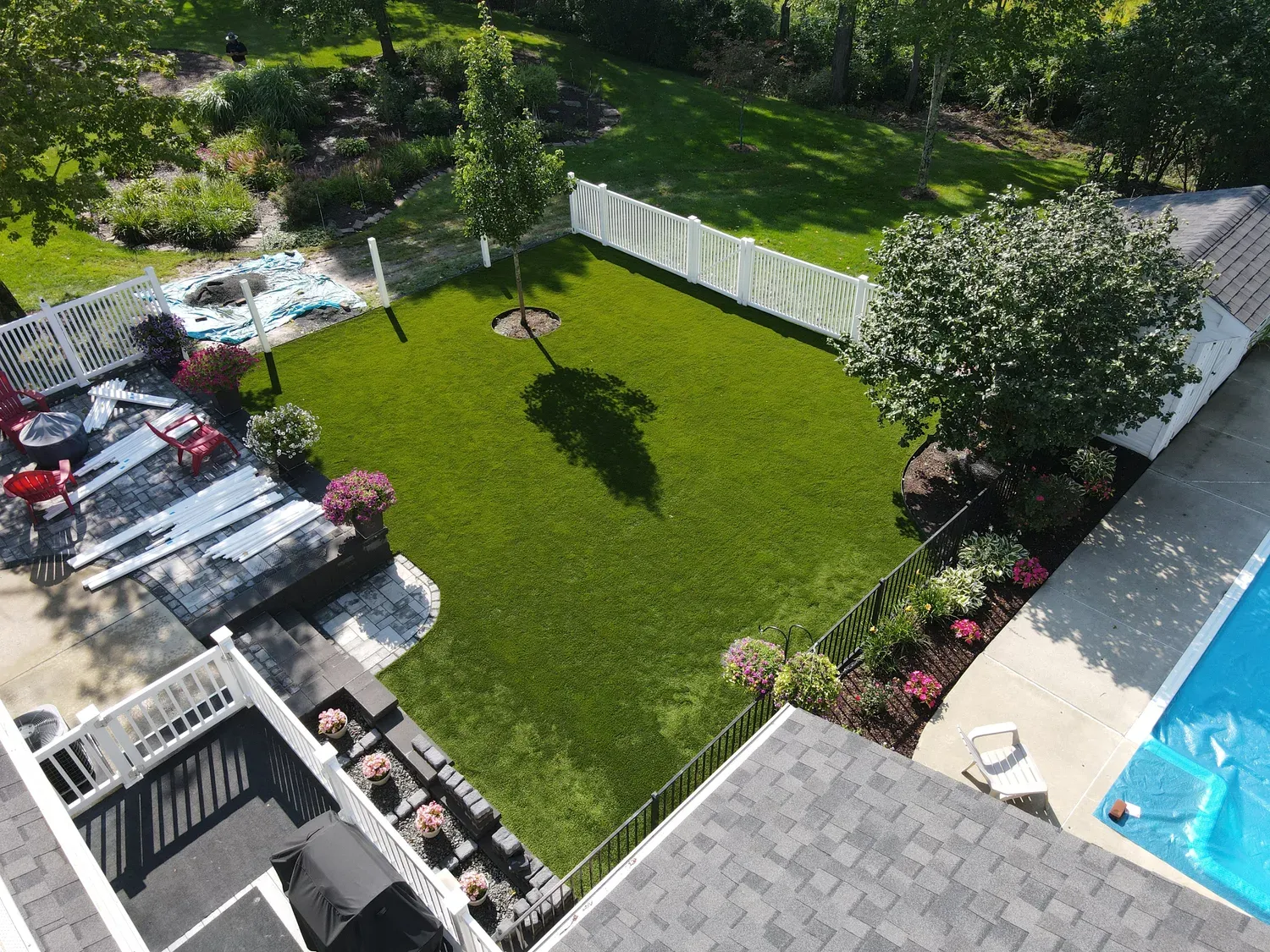 Aerial view of a finished artificial lawn surrounding a mature tree beside a patio, garden border, and white fence