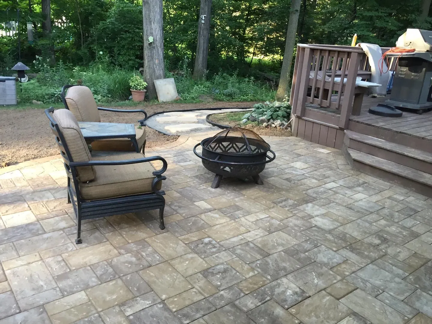 Paver patio in a wooded backyard with cushioned seating arranged around a fire pit