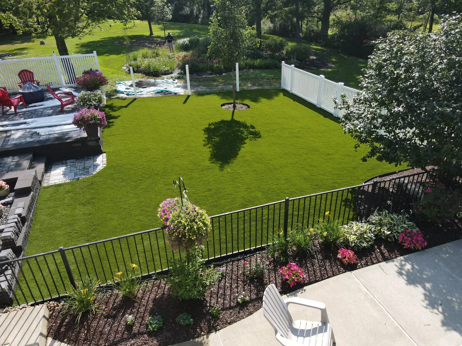 Aerial view of a flat green artificial lawn ringed by a black iron fence, planted borders, and an adjoining paver patio