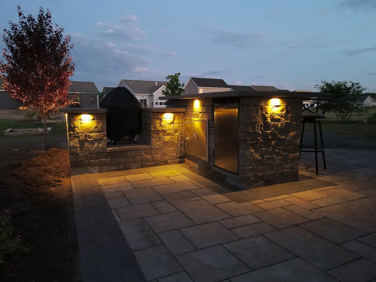 Stone outdoor kitchen island with a stainless door and grill, lit by warm sconces on a paver patio at dusk