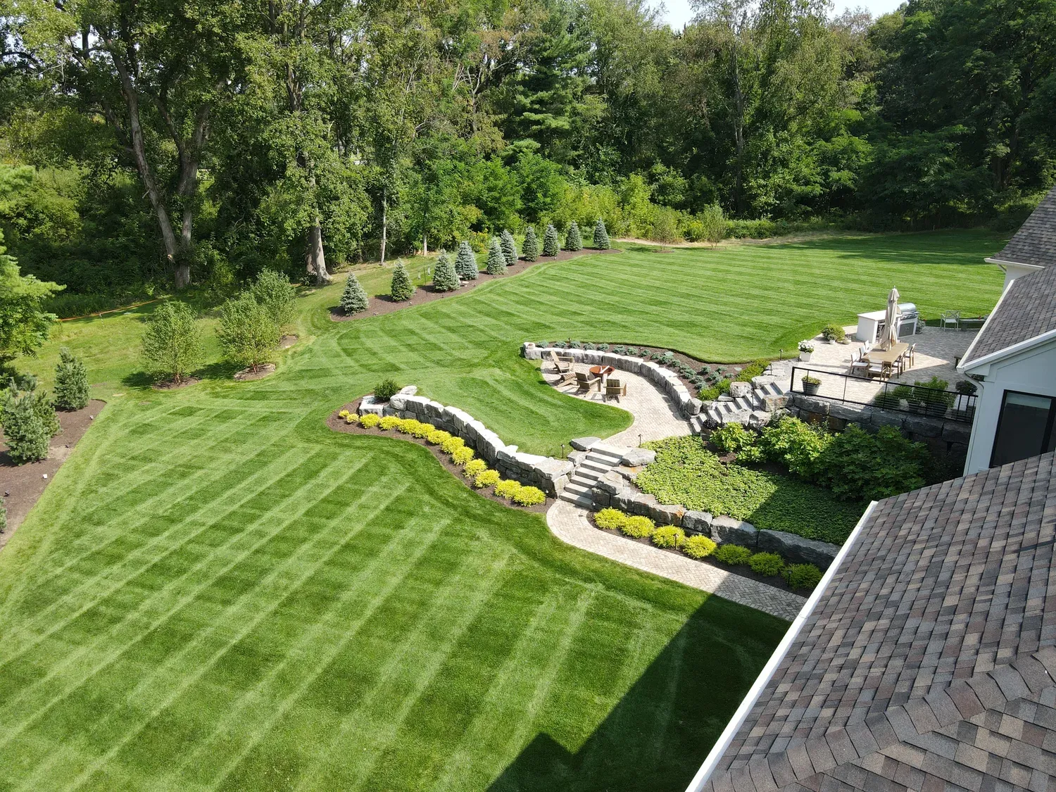 Aerial view of a curved natural stone retaining wall terracing a large striped lawn down to a paver patio and fire pit
