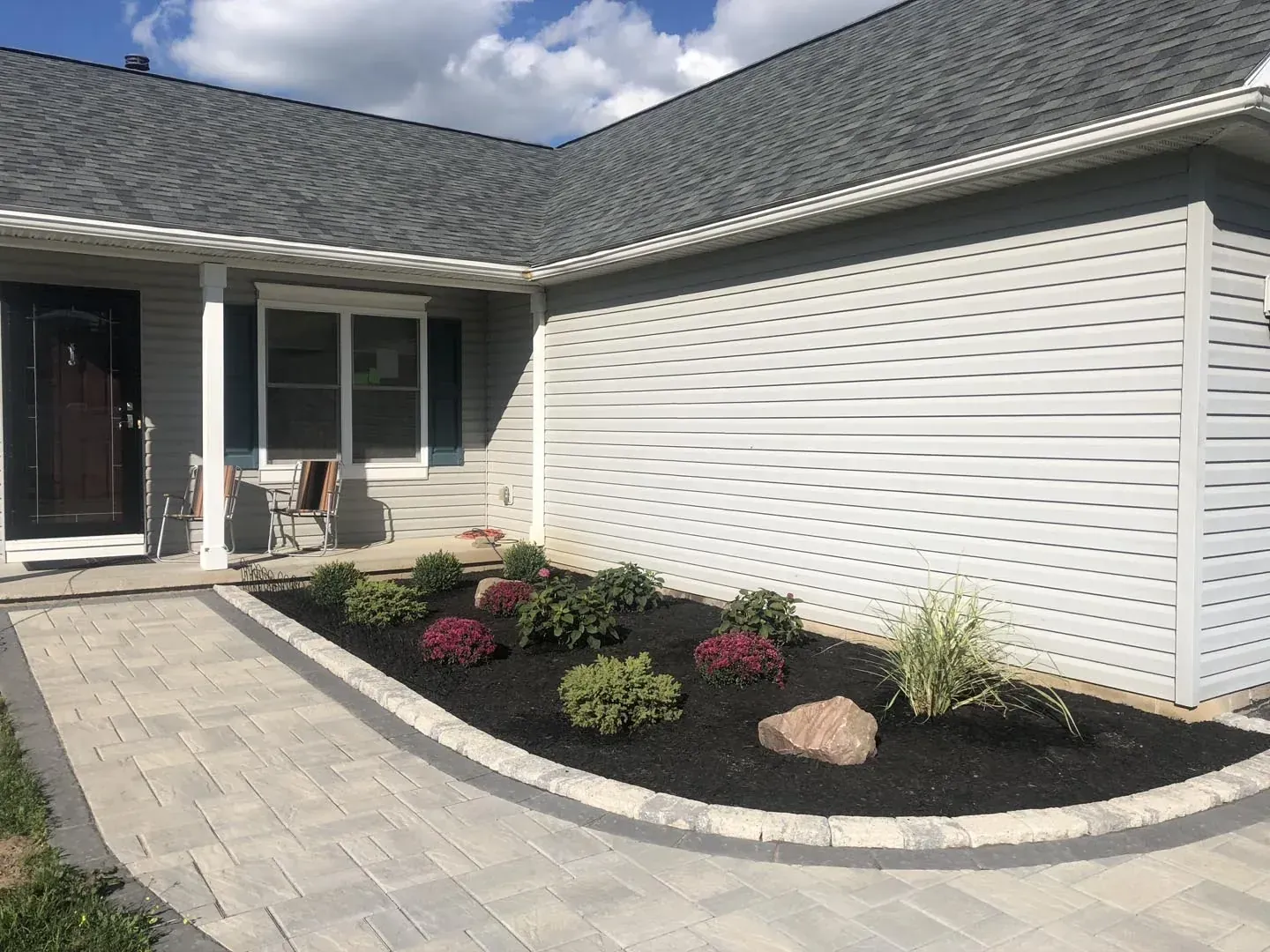 Curved paver walkway with a cobble border leading to a covered front porch past a mulched bed of mums and ornamental grass